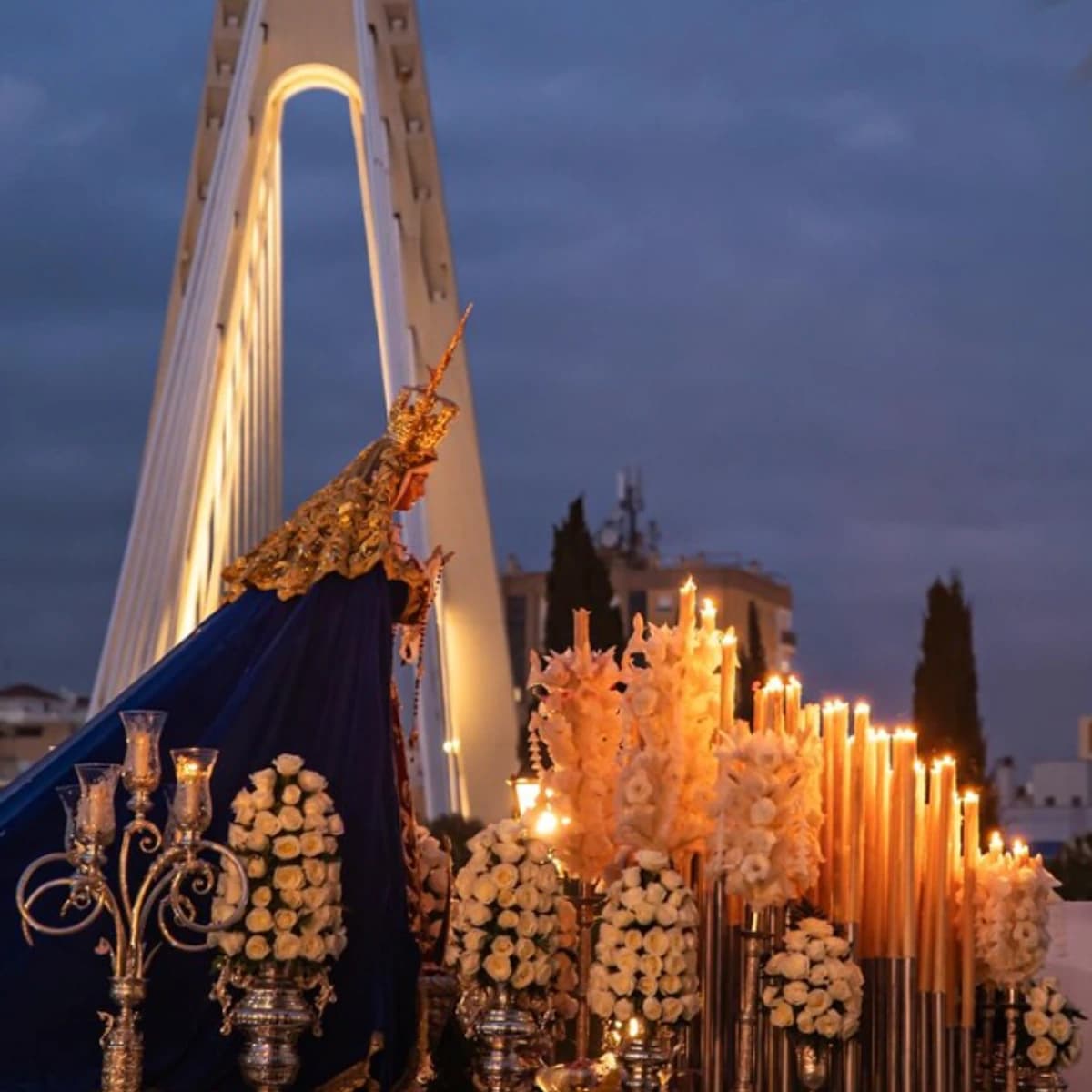 Procesión de Nuestra Señora de la Amargura en Marbella durante el Sábado de Pasión, con el trono de la Virgen iluminado por cirios y rodeado de flores blancas al atardecer, con el puente del arco al fondo.
