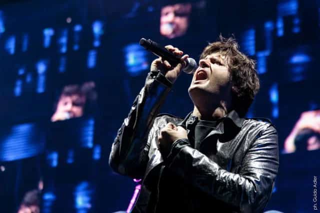 Cantante de rock actuando con micrófono sobre un escenario iluminado en tonos azules, con grandes pantallas al fondo.