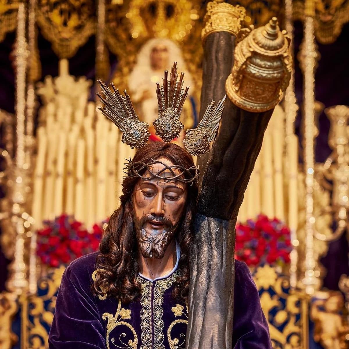 Imagen de Nuestro Padre Jesús Nazareno en Marbella, con túnica morada bordada en oro y la cruz al hombro, en una escena de Semana Santa con fondo de cirios y elementos dorados del trono.