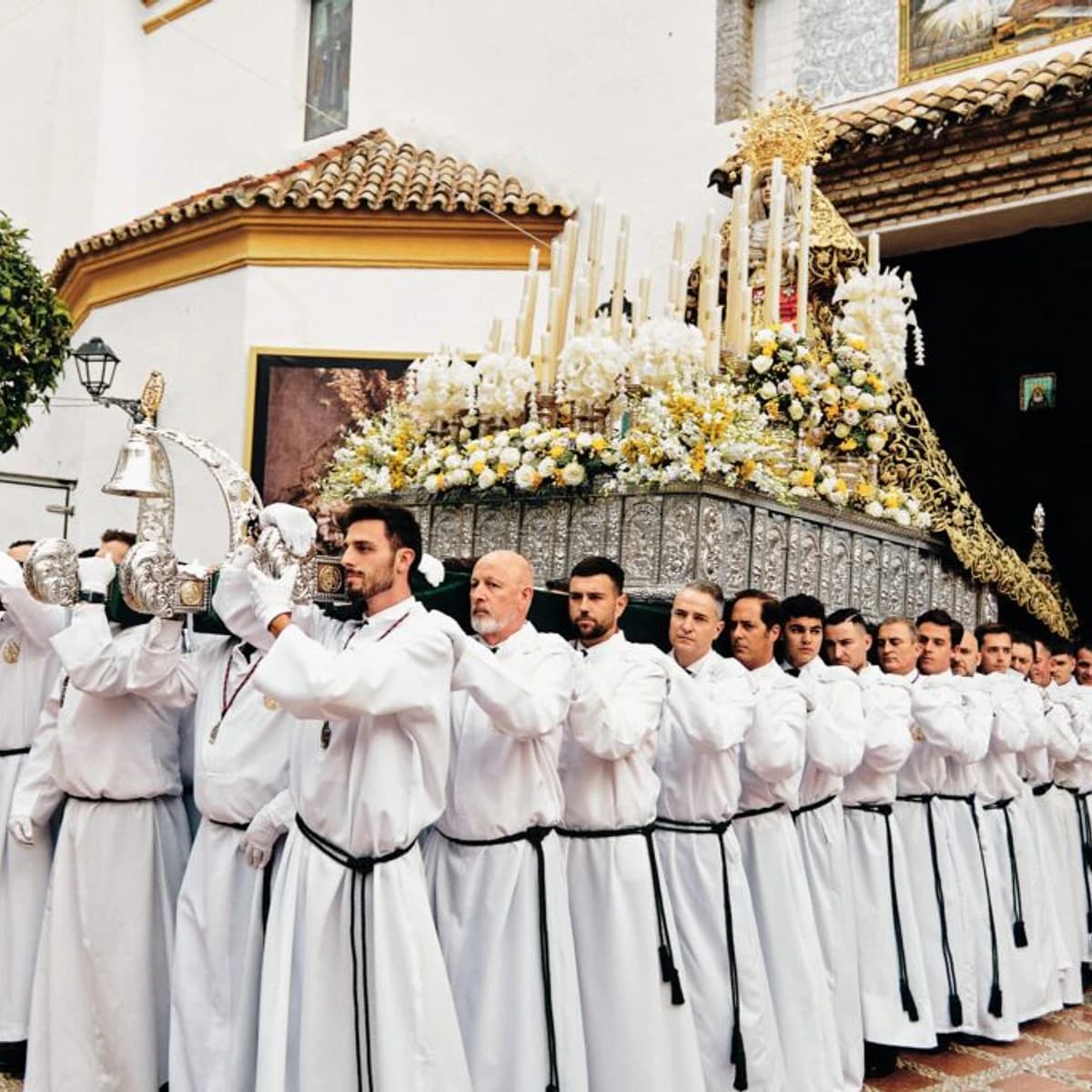 Trono de María Santísima de la Paz y Esperanza de la Cofradía de la Pollinica durante el Domingo de Ramos en Marbella, con portadores vestidos de blanco saliendo del templo entre flores blancas y amarillas.