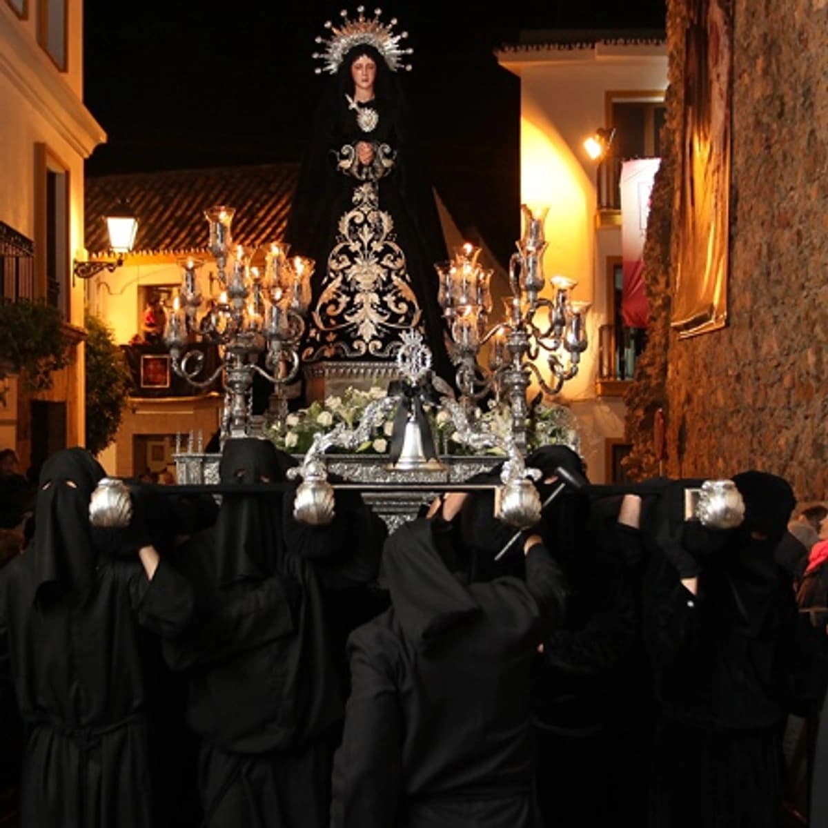 Procesión de Nuestra Señora de la Soledad en Marbella durante el Viernes Santo, con el trono de la Virgen avanzando de noche entre nazarenos de negro, cirios y público en una estrecha calle del centro histórico.