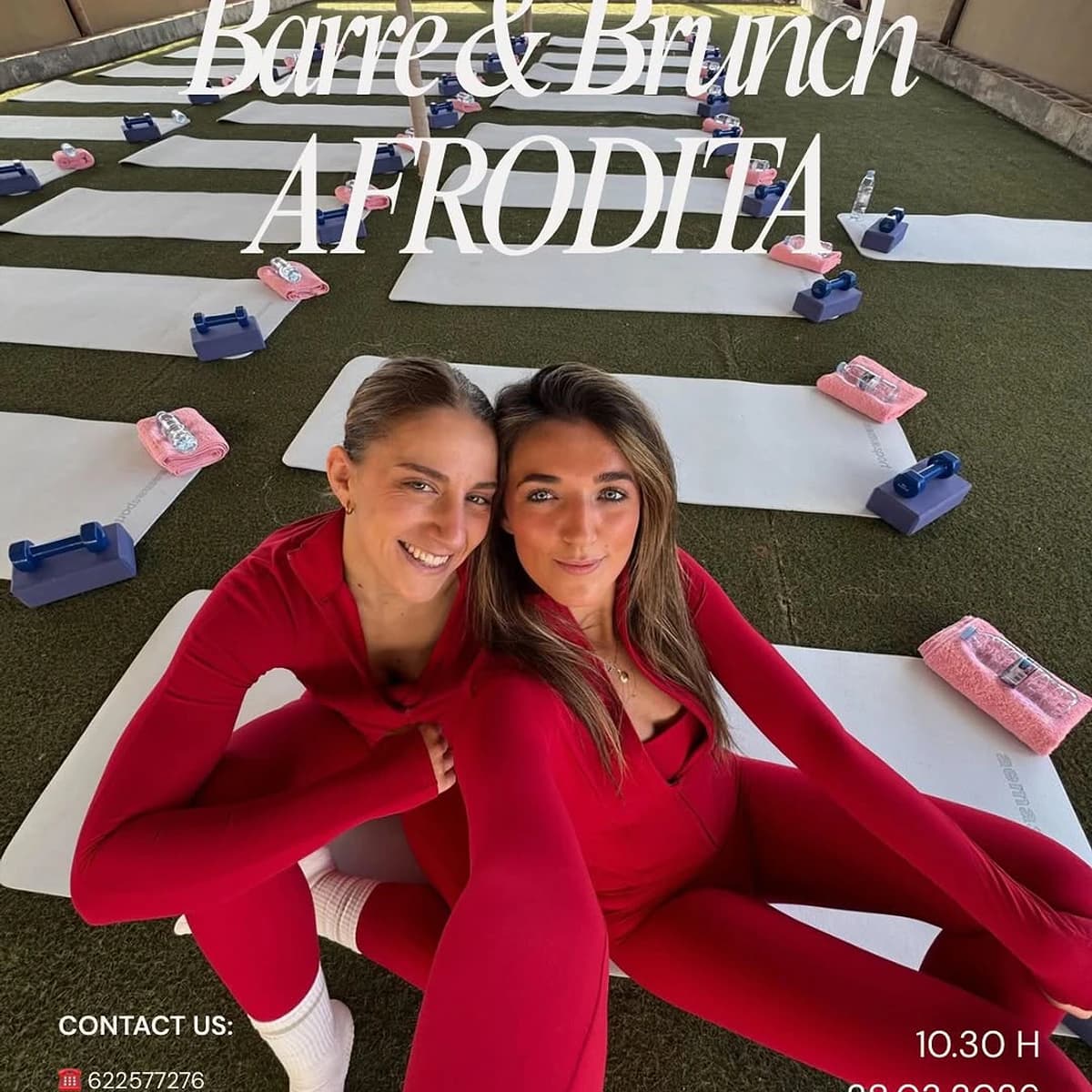 Dos mujeres con ropa deportiva roja sonriendo sobre esterillas de yoga en un evento al aire libre de barre y brunch, con material preparado sobre césped en Afrodíta.