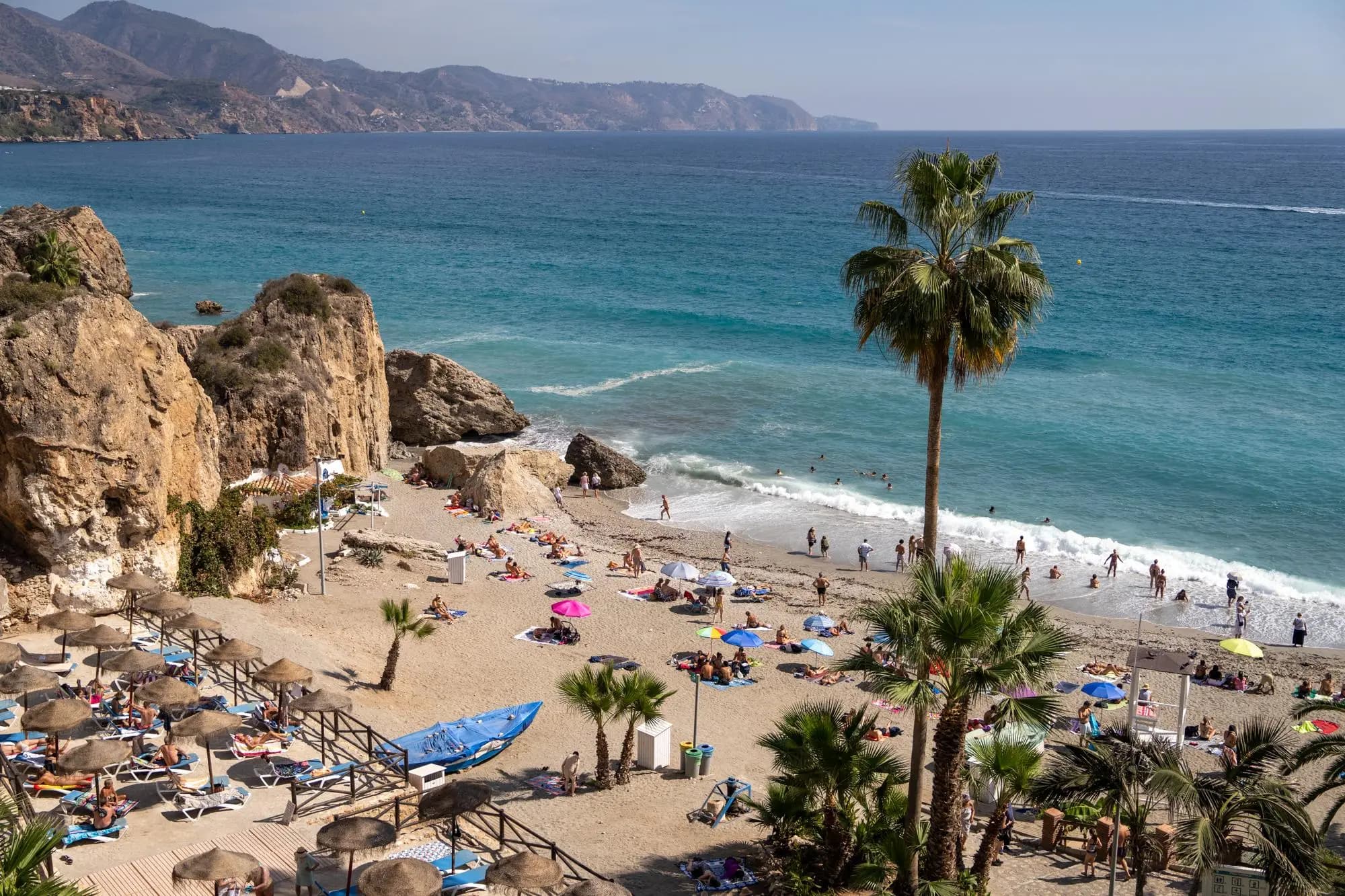 Playa soleada de la Costa del Sol con agua turquesa, palmeras, acantilados rocosos y personas descansando y nadando en la orilla.
