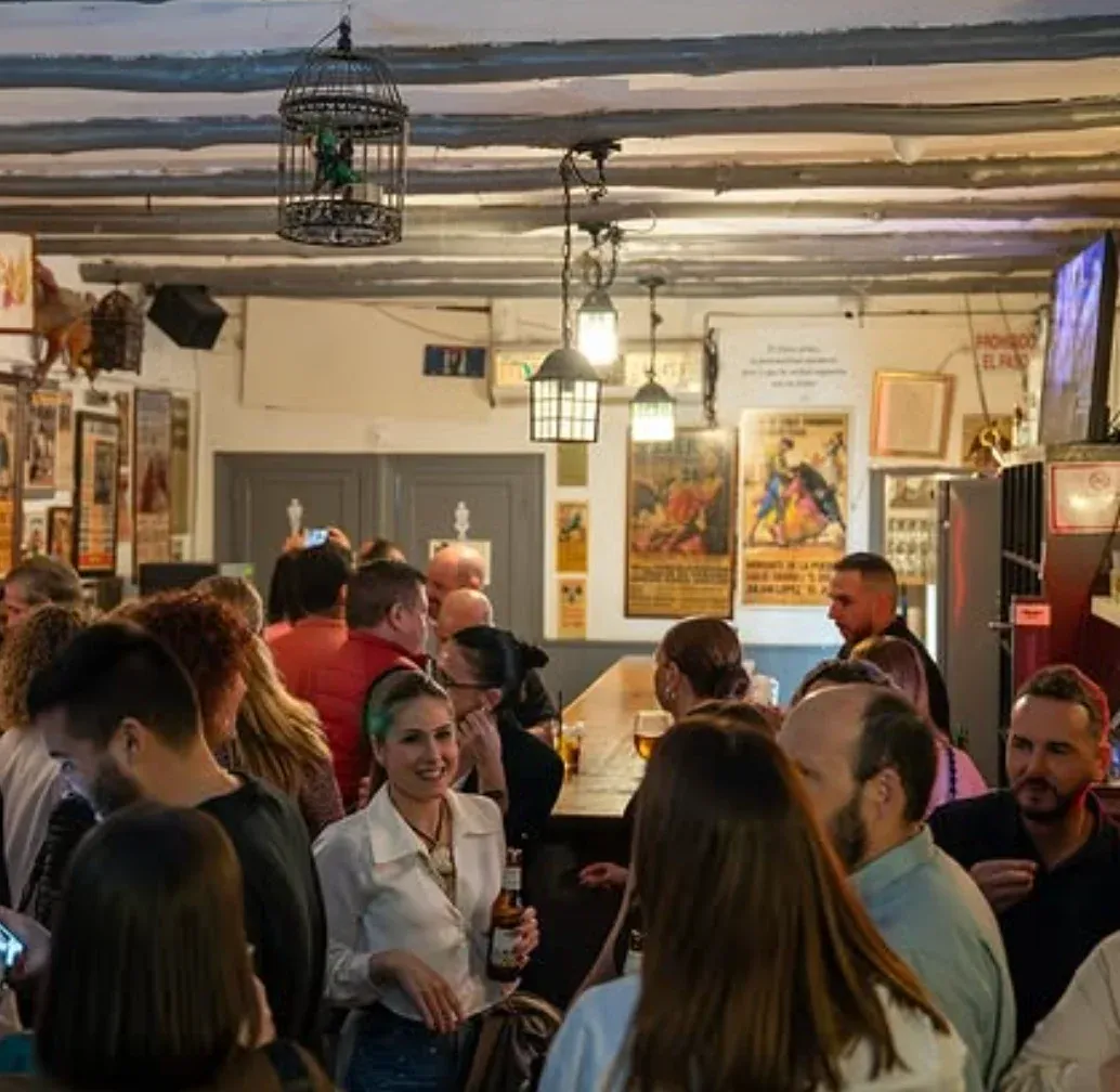 Interior de La Bodeguita de las Pipas con la barra llena de clientes conversando y tomando cerveza, decoración tradicional con carteles taurinos en las paredes y lámparas colgantes en el techo.